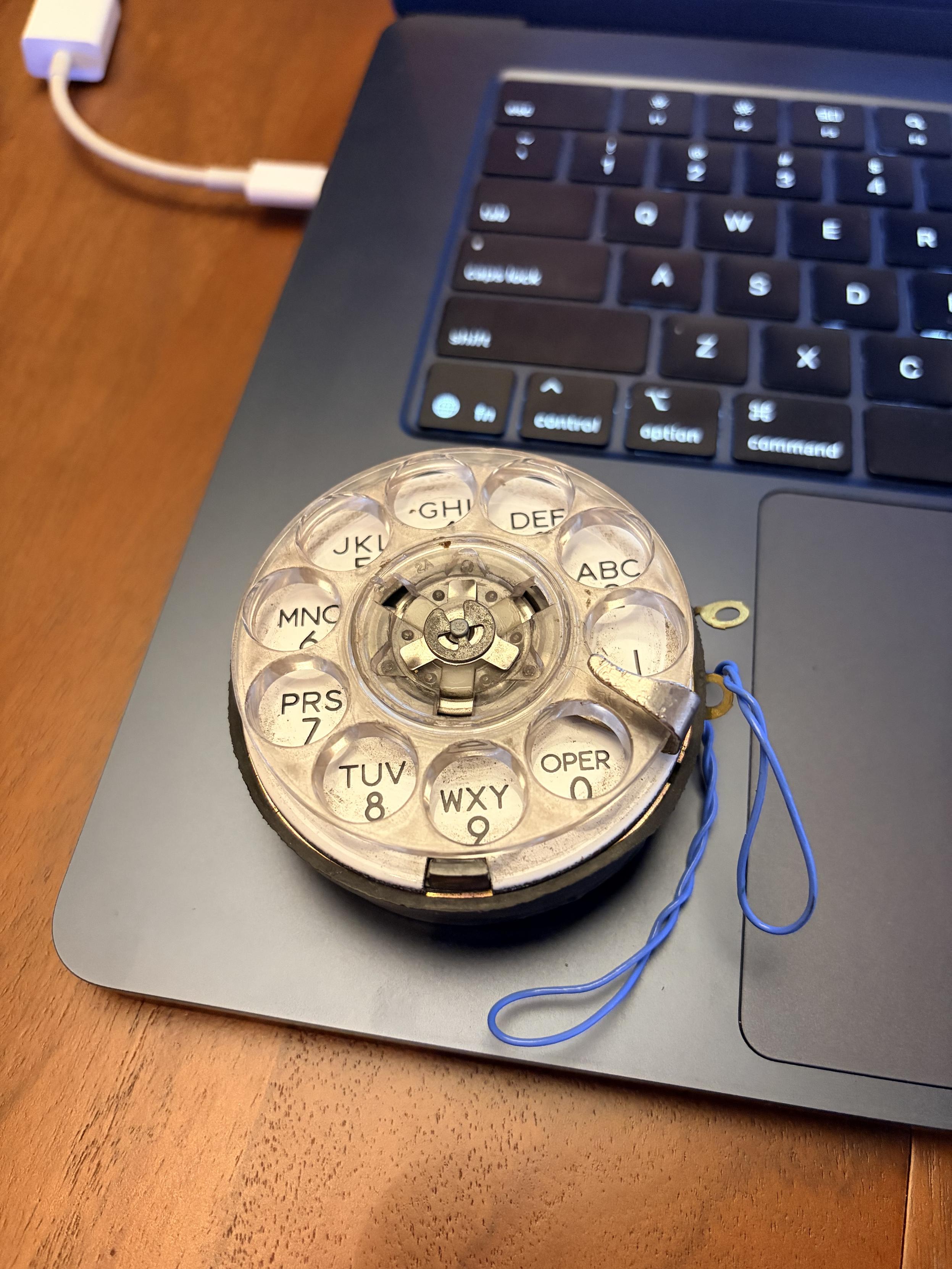 A smoll rotary phone dial with a couple of blue leads coming off it sitting on the corner of my MacBook Air